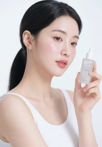 Clear glass bottle with a white dropper cap, labeled "NEEDLY." The model holds the bottle next to her face, showcasing smooth skin.