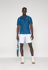 Blå kortermet treningsskjorte med grønne detaljer, hvite shorts, holder en tennisracket og ball, har nettverksstruktur og slim fit.