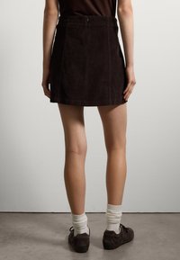 Minifalda de pana marrón con acanalado vertical, presenta un cierre de cremallera en la parte trasera y un diseño ligeramente acampanado. Se lleva con calcetines blancos y zapatos marrones.