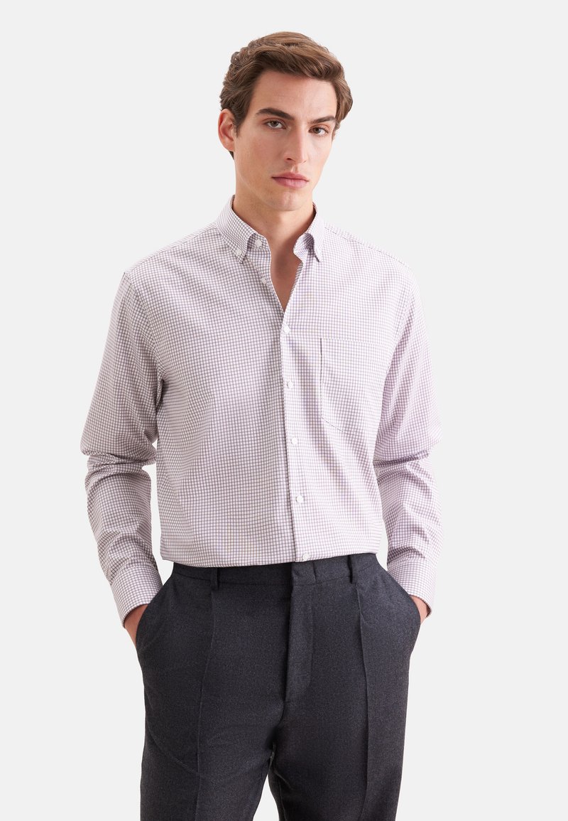 Homme portant une chemise à carreaux claire boutonnée rentrée dans un pantalon sombre sur mesure, les mains dans les poches, debout devant un fond uni.