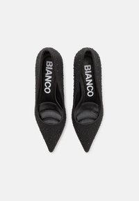 Svarta spetsiga högklackade skor med texturerad ovandel som har små dekorativa pärlor. Inuti har de en slät yta; varumärket "BIANCO" är synligt.
