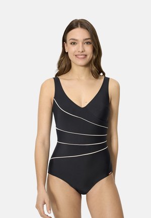 Mujer con cabello castaño medio que lleva un traje de baño negro de una pieza con rayas diagonales blancas, de pie y sonriendo frente a un fondo liso.
