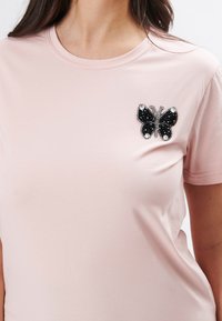 Camiseta de color rosa con una aplicación de mariposa negra y plateada en el lado izquierdo del pecho, con mangas cortas y cuello redondo. Material suave.
