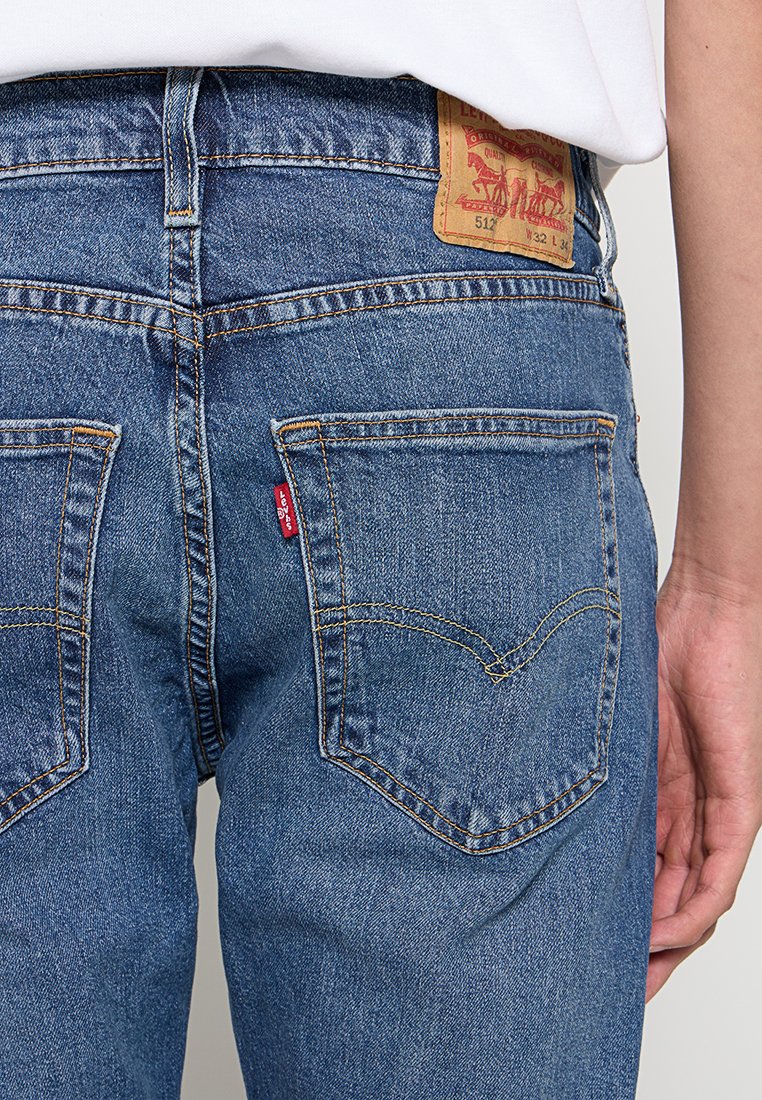 Blaue Jeans aus Denim mit tapered Fit, ausgestattet mit einem roten Etikett und markanten Gesäßtaschen sowie einem beigen Leder-Brandlabel, das an der Taille angebracht ist.