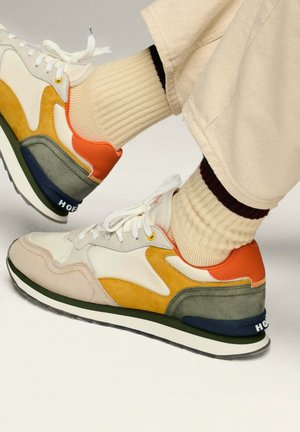 Zapatillas deportivas coloridas que combinan diferentes materiales: cuero beige, acentos de ante naranja y amarillo, tacón gris, y una suela en blanco y negro.