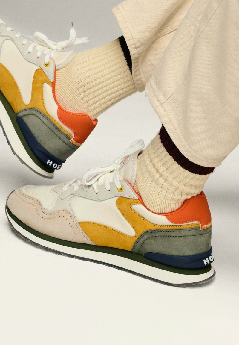 Zapatillas deportivas coloridas que combinan diferentes materiales: cuero beige, acentos de ante naranja y amarillo, tacón gris, y una suela en blanco y negro.