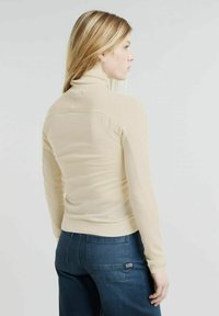 Maglione beige a maniche lunghe con colletto a dolcevita realizzato in tessuto morbido, caratterizzato da un design aderente e texture a coste, abbinato a jeans blu scuro.