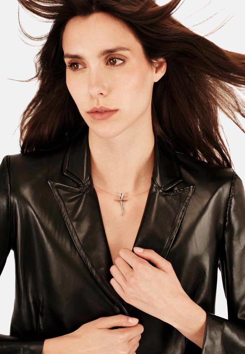 Sølvvedhæng med en slank, forlænget design båret over en sort læderjakke. Modellens hænder griber om jakkens krave.