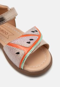 Sandalias brillantes en rosa y naranja con un diseño de rodaja de sandía, que cuentan con acentos de semillas negras y una correa metálica, sobre una suela beige.