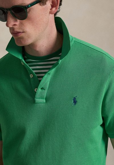 Νέος άνδρας που φοράει πράσινο πόλο με σηκωμένο γιακά πάνω από πράσινη και λευκή ριγέ μπλούζα, και σκούρα γυαλιά ηλίου, που κοιτά προς τα αριστερά.