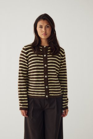 Junge Frau trägt eine braun-creme gestreifte, geknöpfte Strickjacke und dunkle weite Hosen und steht vor einem schlichten hellen Hintergrund.