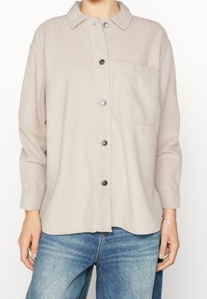 Béžová manžestrová košile na knoflíky s náprsní kapsou, nošená s modrými džínami, dlouhými rukávy a stříbrným náramkem s přívěsky na pravém zápěstí.