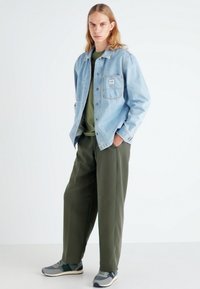 Veste en jean bleu clair sur une chemise verte foncée, assortie à un pantalon ample vert olive et des baskets grises. La tenue est décontractée et relax.