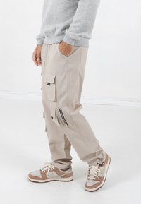 Beige Cargo-Hosen mit mehreren Taschen und Reißverschluss-Akzenten, kombiniert mit hellgrauen Turnschuhen mit braunen und cremefarbenen Details. Weiche Stofftextur.