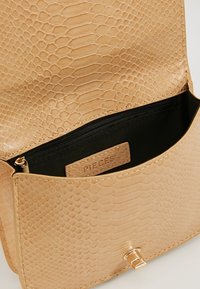 Béžová kabelka se vzorem hadí kůže, magnetickým zapínáním, černou kapsou na zip uvnitř a značkovou visačkou. Hladký povrch a lesklé detaily.