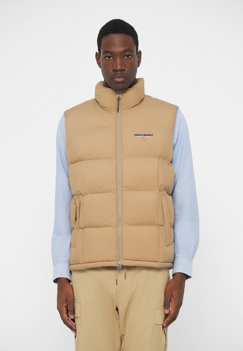 Gilet imbottito beige con colletto alto, zip anteriore, tasche laterali e logo ricamato. Indossato sopra una camicia a maniche lunghe azzurro chiaro.