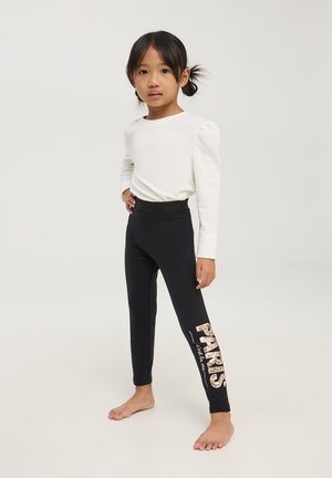 Leggings neri con la scritta "PARIS" stampata in rosa metallizzato sulla gamba sinistra. Abbinati a una maglietta bianca a maniche lunghe con spalle a palloncino.