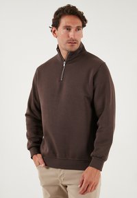 Bruine sweatshirt met rits en lange mouwen, met ribbelmanchetten en een gladde, gestructureerde stof; gecombineerd met lichtbeige broek.