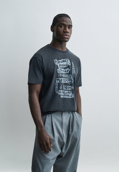 Black man standing with hands in pockets wearing a black graphic T-shirt and loose gray pleated pants against a plain light background.
