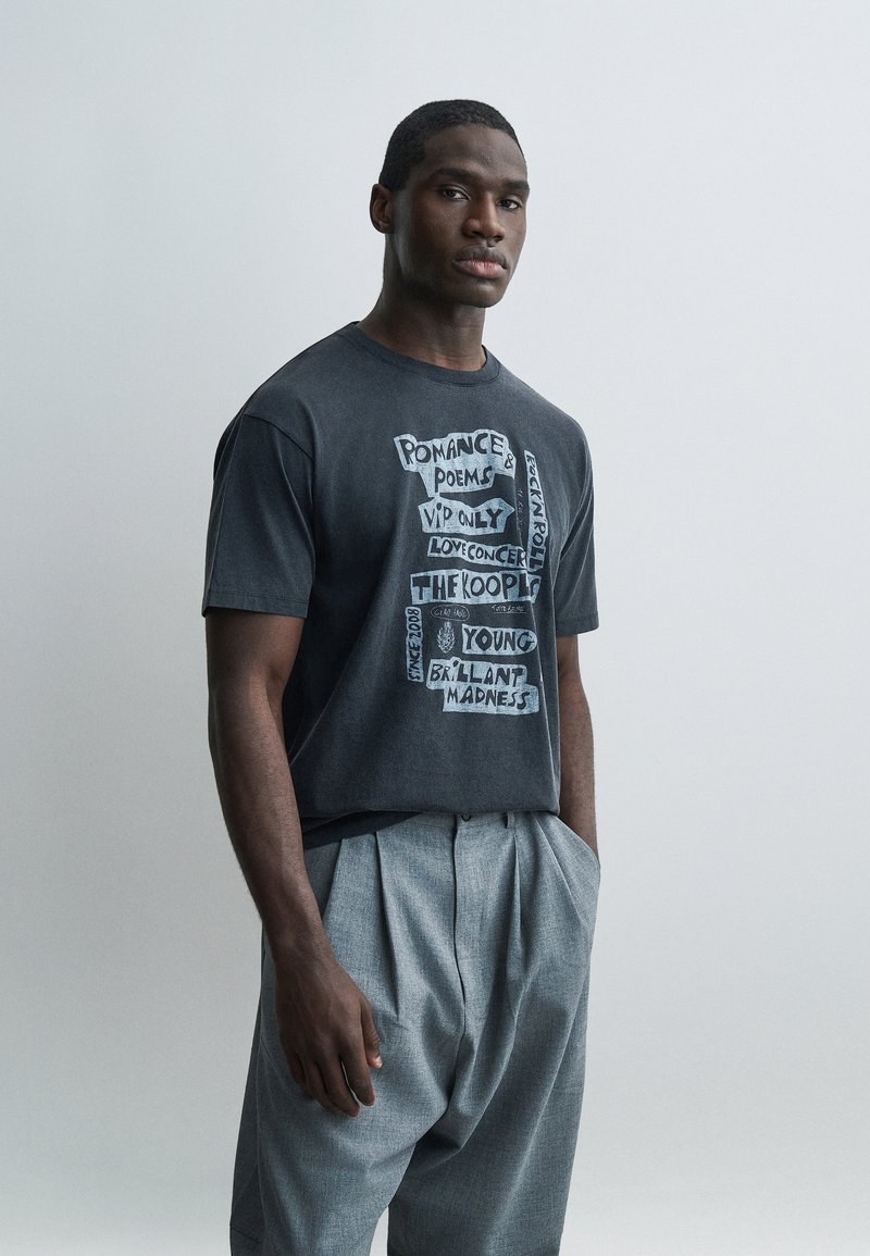 Μαύρος άνδρας στέκεται με τα χέρια στις τσέπες, φορώντας μαύρο μπλουζάκι με σχέδιο και χαλαρό γκρι πλισέ παντελόνι, πάνω σε απλό ανοιχτό φόντο.