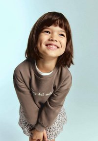 Sweatshirt maro cu text alb "Mic dar sălbatic", guler crem; asociat cu o fustă cu imprimeu leopard. Fundal bleu deschis.