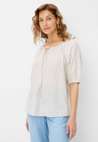 Lys beige bluse med lodrette striber, rund halsudskæring og nøglehulsdetalje. Løse ærmer med elastiske manchetter, parret med lyseblå jeans.