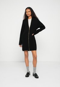 Cardigã de malha preta canelada com fecho de correr, mangas compridas e bolsos frontais. Usado por cima de uma blusa clara e combinado com meias cinzentas e sapatos pretos.