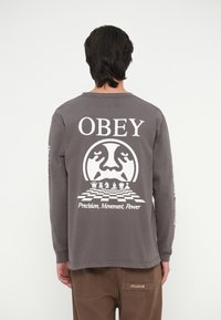 Siva majica dugih rukava s bijelim grafikama na leđima: "OBEY", dizajn šahovske ploče i tekst "Preciznost, Kretanje, Snaga". Mekani materijal, opušteni kroj.