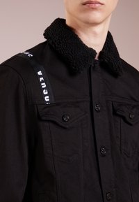 Giacca di denim nero con colletto in pelliccia nera testurizzata, dotata di due tasche sul petto, chiusura con bottoni e cinghie con marchio sulla spalla.