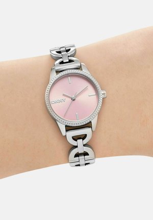 Silberne DKNY-Armbanduhr mit pinkem Zifferblatt und minimalistischen Markierungen auf einem gliedernden Metallarmband, getragen am Handgelenk einer Person.