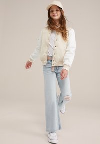 Bež in bela bomber jakna, bela majica, svetlo modri raztrgani kavbojke in bele superge z rdečimi detajli. Model nosi bež kapo.