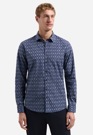 Man draagt een strakke, lange mouw, blauw shirt met een diamantpatroon en zwarte broek, staand tegen een effen witte achtergrond.