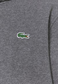 Logo de crocodile vert brodé avec une bouche rouge sur un tissu en maille gris, positionné près du centre du vêtement.