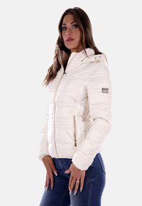 Giacca bianca trapuntata con cappuccio, dotata di tasche con zip e dettagli in metallo color oro, realizzata in un materiale lisco e lucido.