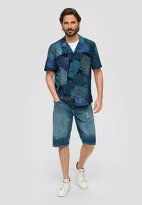 Korte mouwen navy shirt met abstracte blauwe en turquoise patronen; gecombineerd met distressed denim shorts en witte sneakers.