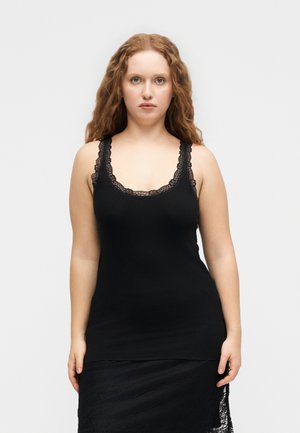 Woman with long curly hair wearing a black ribbed tank top with lace trim and a black lace skirt against a plain light background.