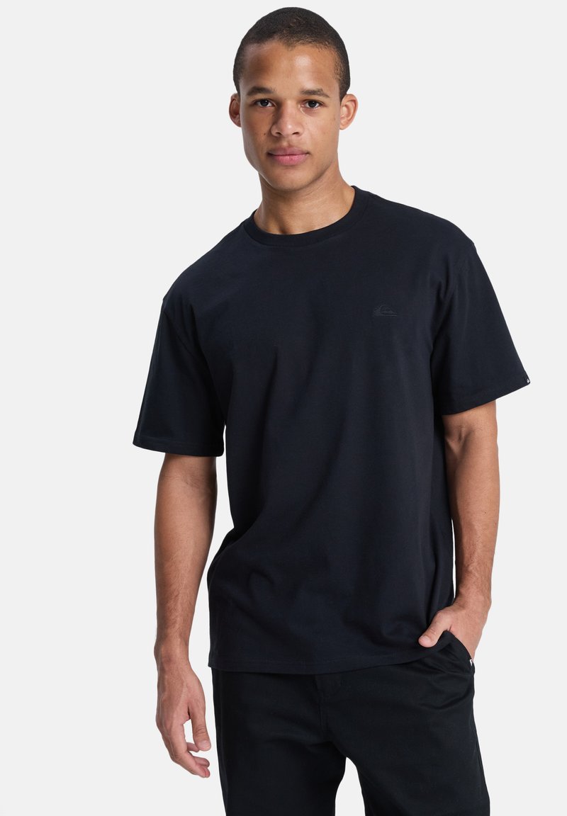 Jeune homme debout, une main dans la poche, portant un t-shirt noir uni et un pantalon noir sur fond blanc.