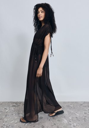 Μαύρο, διάφανο μάξι φόρεμα με κοντά μανίκια, σούρωμα στη μέση και διακοσμητικά δέσιμο. Φορεμένο με μαύρα σανδάλια σε υφή πέτρινης επιφάνειας.