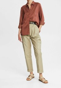 Persoon in een roestkleurig longsleeve shirt, beige cargobroek met zakken, zwarte riem en beige sandalen, staand tegen een witte achtergrond.