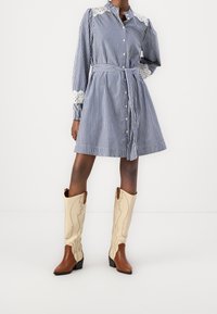 Randig blå skjortklänning med knappstängning, spetsdekorerade ärmar och knytning i midjan; ihop med bruna och krämfärgade cowboystövlar med detaljer.