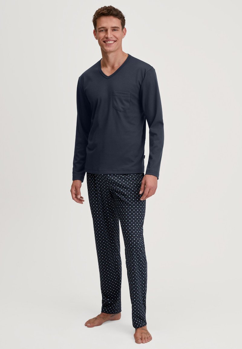 Maglia a maniche lunghe blu navy con scollatura a V e taschino sul petto, abbinata a pantaloni da pigiama a fantasia con pois bianchi su uno sfondo scuro.