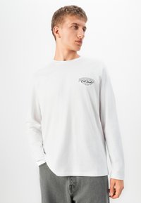 Jaunas vyras vilki baltą ilgomis rankovėmis Jack & Jones marškinėlį ir pilkas kelnes, žiūri į šoną, viena ranka kišenėje.