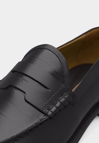 Schwarzer Lederloafer mit strukturierter Oberfläche und einem Pennyriemen-Detail, sichtbaren Nähten und einem hellbraunen Innenfutter.