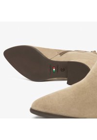 Bottines en suède beige à bout pointu avec un détail de fermeture éclair. La semelle en caoutchouc est plate avec une texture nervurée, arborant un logo et le drapeau italien.