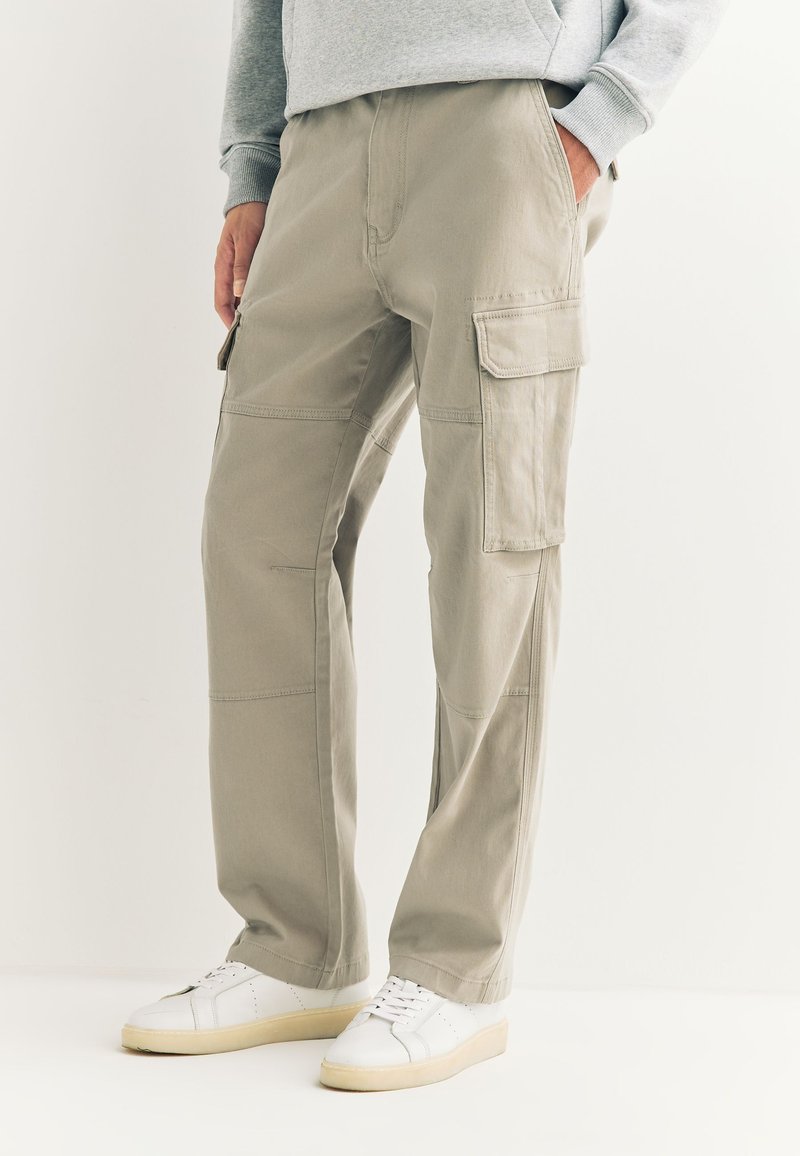 Pantalon cargo beige avec une coupe décontractée, doté de poches latérales, passants pour ceinture et surpiqûres contrastantes, associé à des baskets blanches.