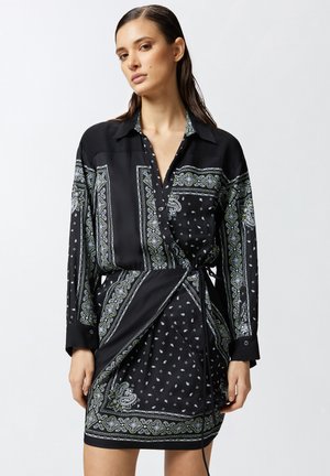 Il vestito in satin nero presenta un colletto, maniche lunghe e intricati motivi paisley verdi e bianchi. Include un design avvolgente con una cintura da annodare.