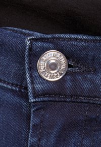 Botón de tela vaquera con texto grabado, color azul oscuro, que presenta una costura de sobrehilado y una línea de costura vertical cerca del botón.