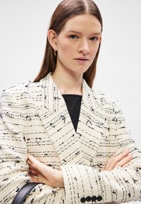 Λευκό σακάκι από tweed με μαύρες υφασμάτινες ρίγες, με γιακά τύπου notch, και τρία κουμπιά σε κάθε μανίκι. Φοριέται πάνω από ένα μαύρο τοπ.