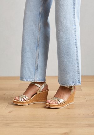 Fødder iført gyldne kilehælssandaler med stropper, kombineret med lyseblå jeans med lige ben, der står på trægulv.