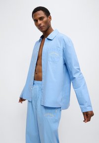 Ανοιχτό γαλάζιο πουκάμισο με ρίγες και τσέπη με χρυσά γράμματα "Calvin Klein", συνδυασμένο με παντελόνι ίδιου σχεδίου σε χαλαρή εφαρμογή.